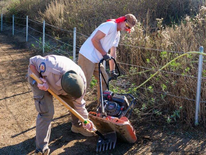 Two men work on fixing a trail, one is bent over scraping dirt with a tool off of a machine held by the other.